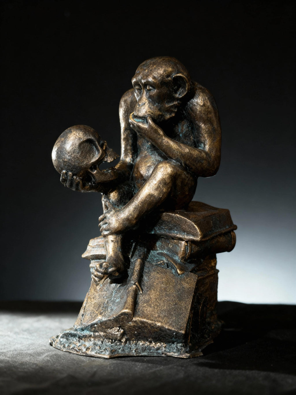 May include: Bronze sculpture of a monkey seated atop books, holding a skull. The monkey is in a contemplative pose, with its hand near its mouth. The sculpture has a dark, aged finish, and the details of the monkey and skull are clearly visible.