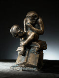 May include: Bronze sculpture of a monkey seated atop books, holding a skull. The monkey is in a contemplative pose, with its hand near its mouth. The sculpture has a dark, aged finish, and the details of the monkey and skull are clearly visible.
